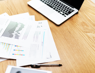 木目のテーブルにノートパソコンとグラフが書かれた資料が置かれた図