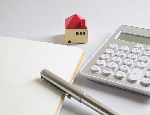 白い机に電卓と家の型をした置物、ノートとペンが置かれた図